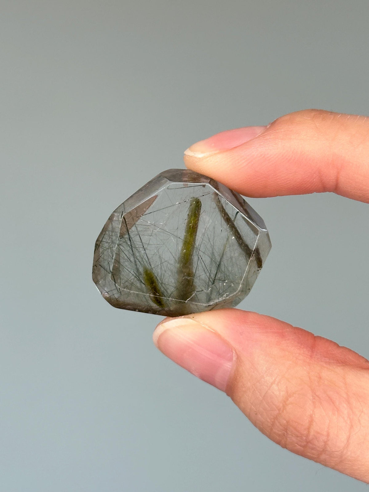 May include: A faceted, clear quartz crystal with green and black inclusions. The crystal is held between two fingers against a gray background. The inclusions appear as needle-like structures within the transparent stone.