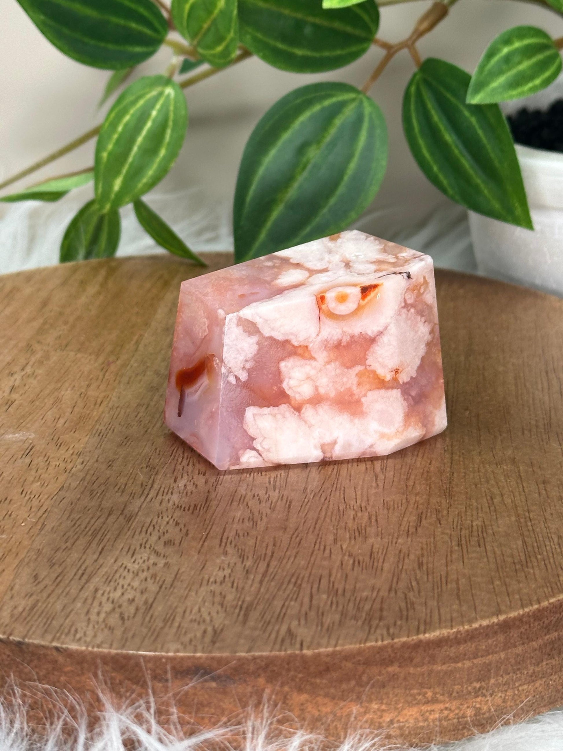 May include: A faceted, rectangular crystal in shades of pink and white, with orange and brown inclusions. The crystal sits on a round, wooden tray with a light brown finish. Green leaves are visible in the background.