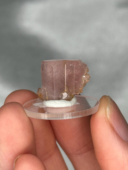 May include: A light pink crystal specimen with a rectangular shape and visible facets. The crystal is mounted on a clear, circular base with a white adhesive. The background is a soft, neutral color.