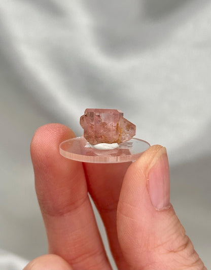 May include: A small, pink crystal specimen is displayed on a clear, circular stand. The crystal has a rough, natural texture and a translucent appearance. The stand is held by a person's fingers against a soft, white background.