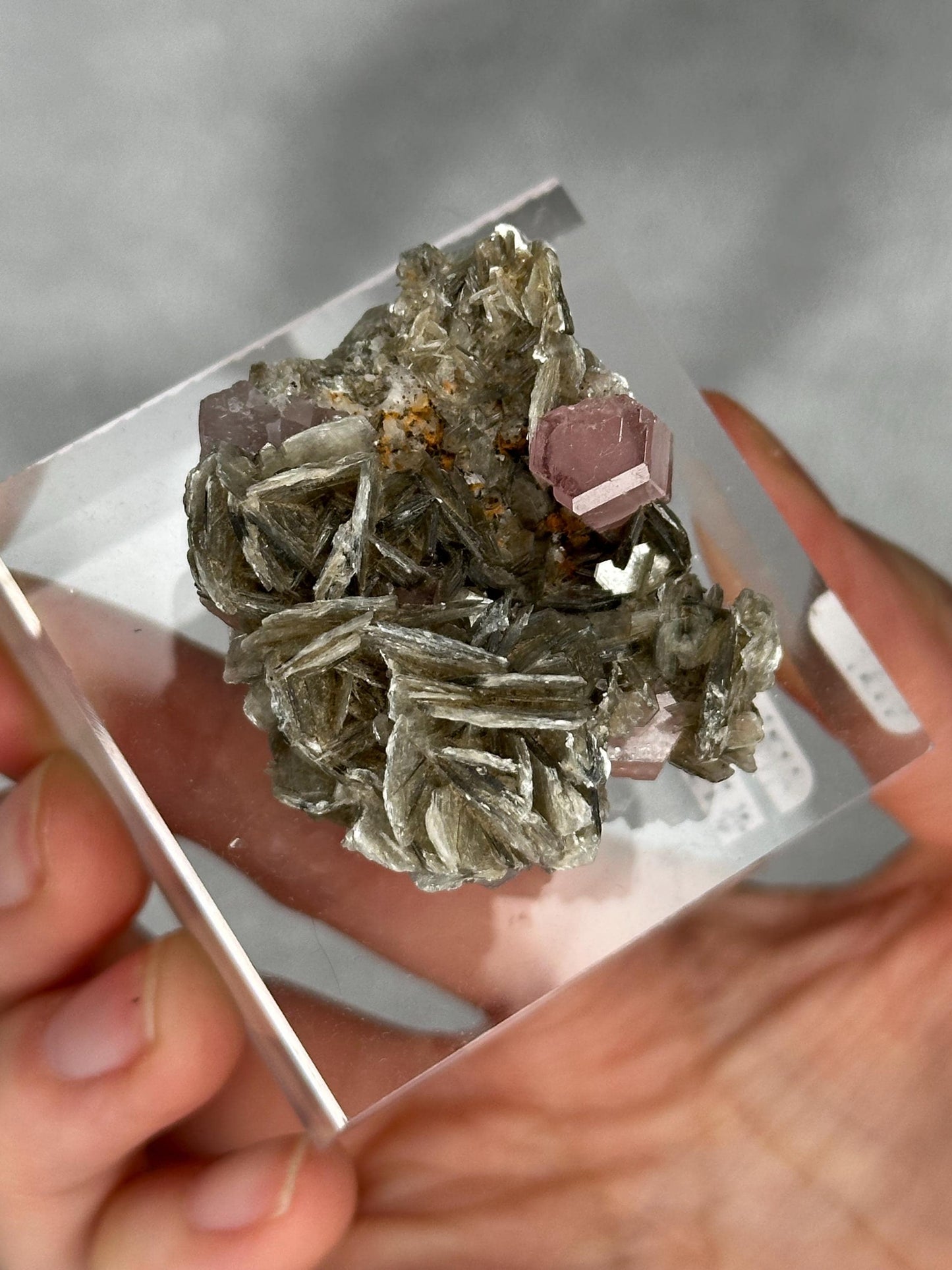 May include: A mineral specimen featuring a cluster of gray, layered crystals with a few small, pink, hexagonal crystals. The mineral is encased in a clear acrylic display case, held by a hand.