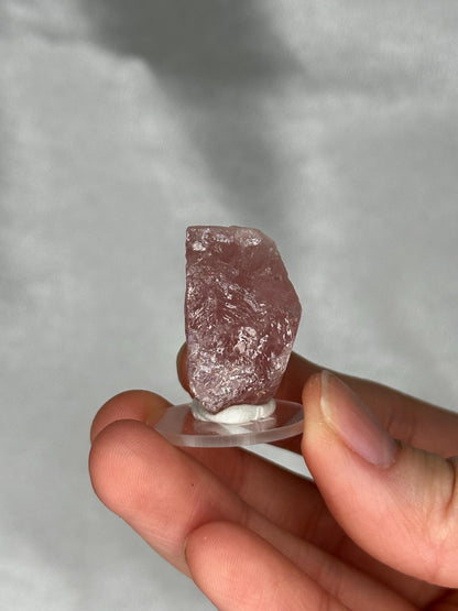 May include: A rough-cut, pink crystal specimen mounted on a clear, circular base. The crystal has a textured surface and varying shades of pink. The base is held by a hand against a soft, neutral background.