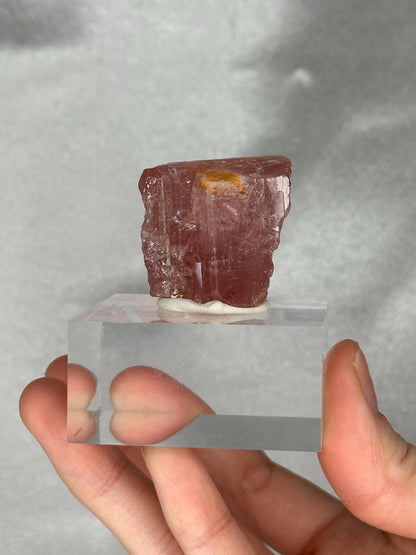 May include: A pink tourmaline crystal specimen, mounted on a clear acrylic base. The crystal has a rough, textured surface and a rectangular shape. The base is held by a hand.