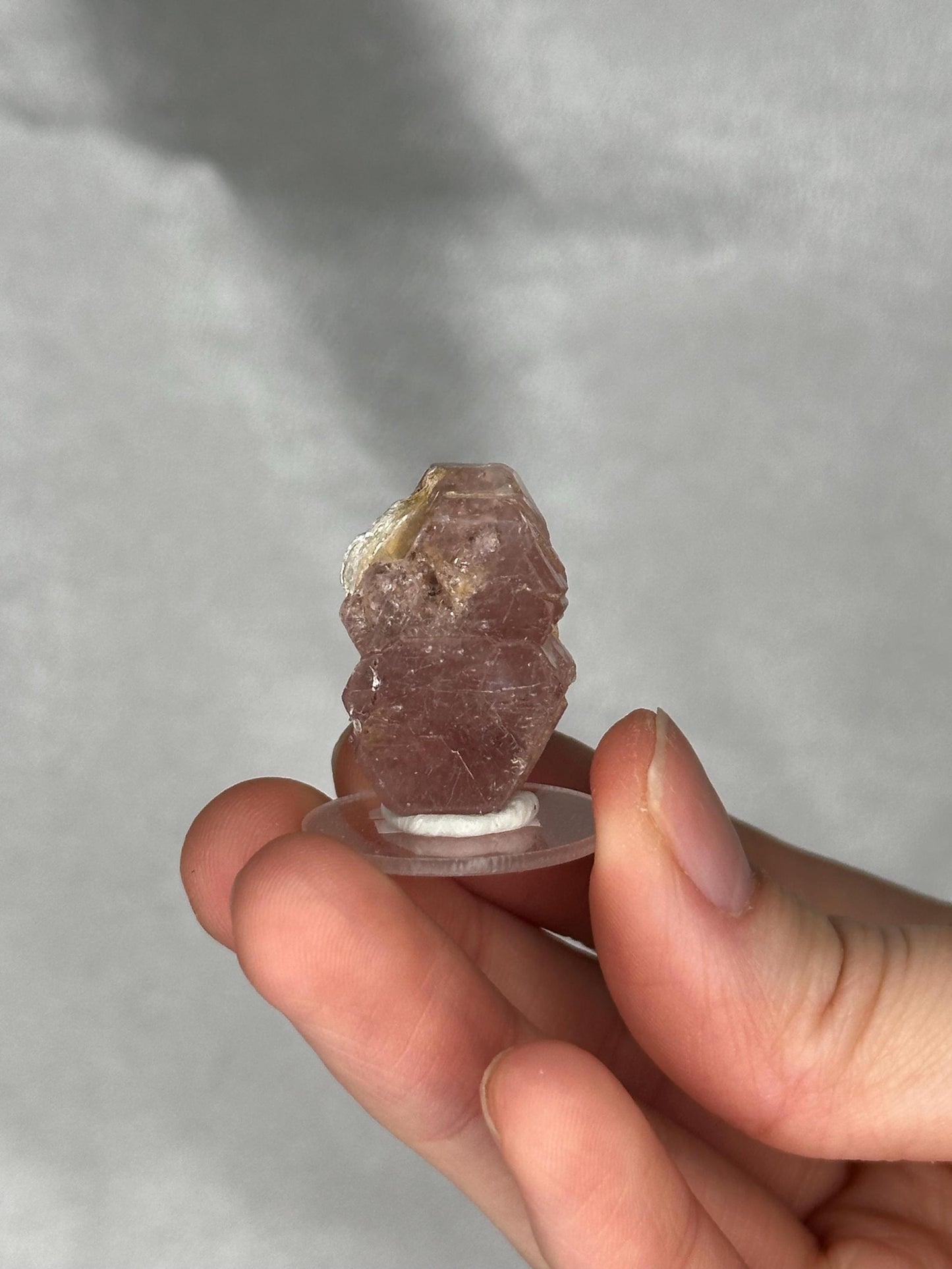 May include: A small, rough-cut, pinkish-purple crystal specimen. The crystal is mounted on a clear, circular base with a white adhesive. The crystal has a natural, unpolished appearance, with visible texture and variations in color.