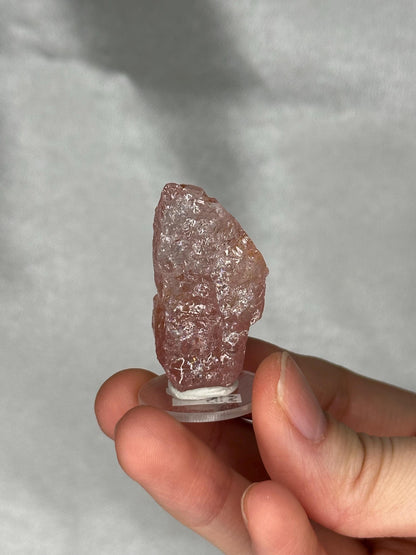 May include: A rough, pink crystal specimen, held on a clear, circular stand. The crystal has a textured surface and varying shades of pink. The image is well-lit, highlighting the natural details of the mineral.