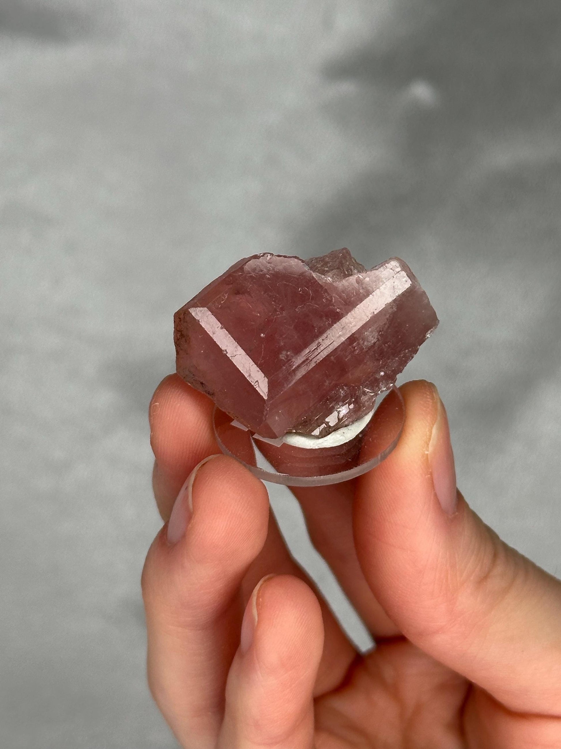 May include: A faceted, pink crystal specimen. The crystal is held on a clear, circular stand. The crystal has a geometric shape with visible facets and a translucent appearance. The background is a solid, neutral gray.