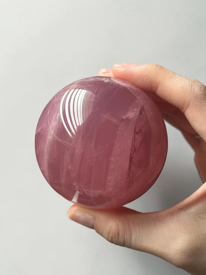May include: A polished, spherical rose quartz crystal with a translucent, pale pink hue. The surface displays natural internal fractures and inclusions, with a bright white light reflection visible on the upper left side of the smooth, rounded stone.